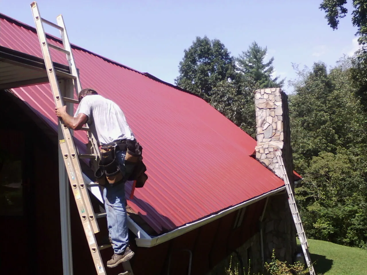 Roofer on a ladder beside a red metal roof and stone chimney — Soffit & Fascia Repair by Roofing Brothers in Kitzmiller